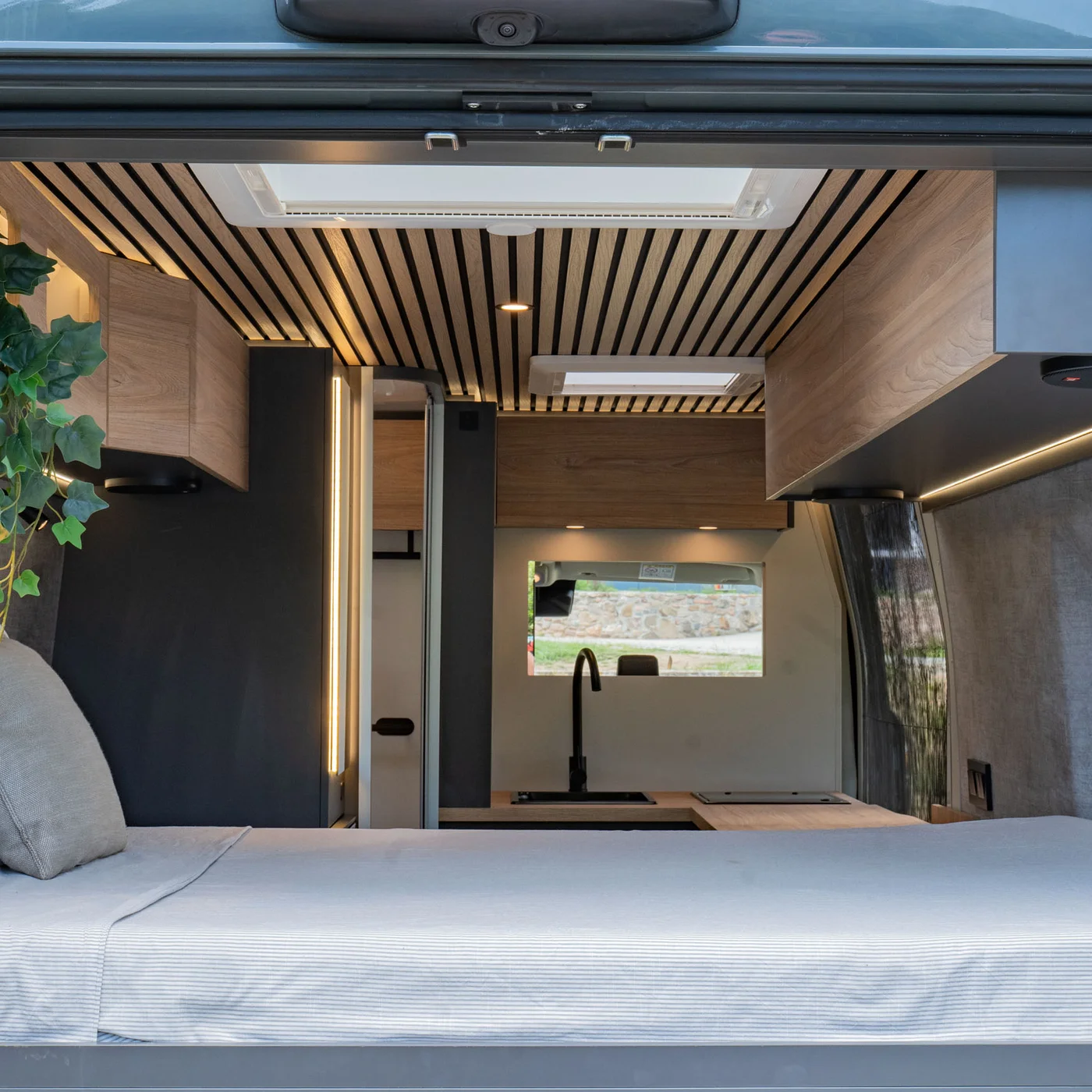 Interno di un camper moderno con letto, armadi in legno e pianta verde.