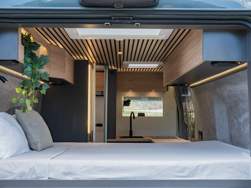 Interno di un camper moderno con letto, armadi in legno e pianta verde.