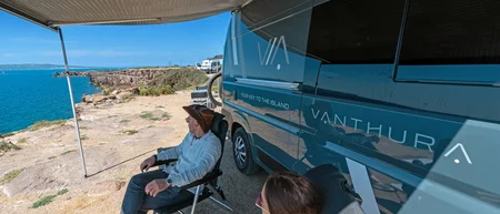 Zwei Personen sitzen neben einem blauen Wohnmobil mit Meerblick