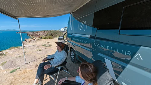 Zwei Personen sitzen neben einem blauen Wohnmobil mit Meerblick