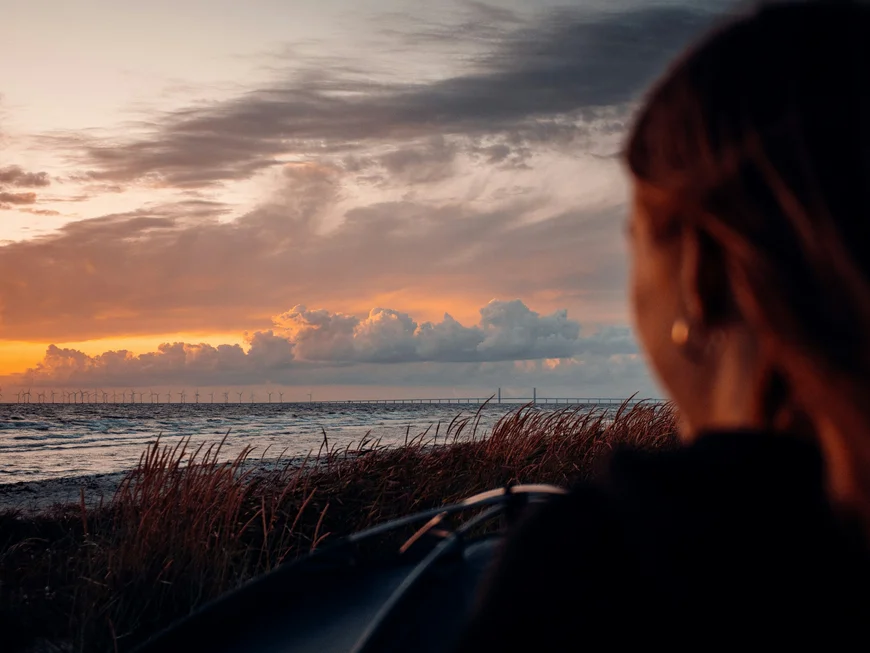 Donna che osserva il tramonto sull'oceano con cielo nuvoloso e vento sulle erbe
