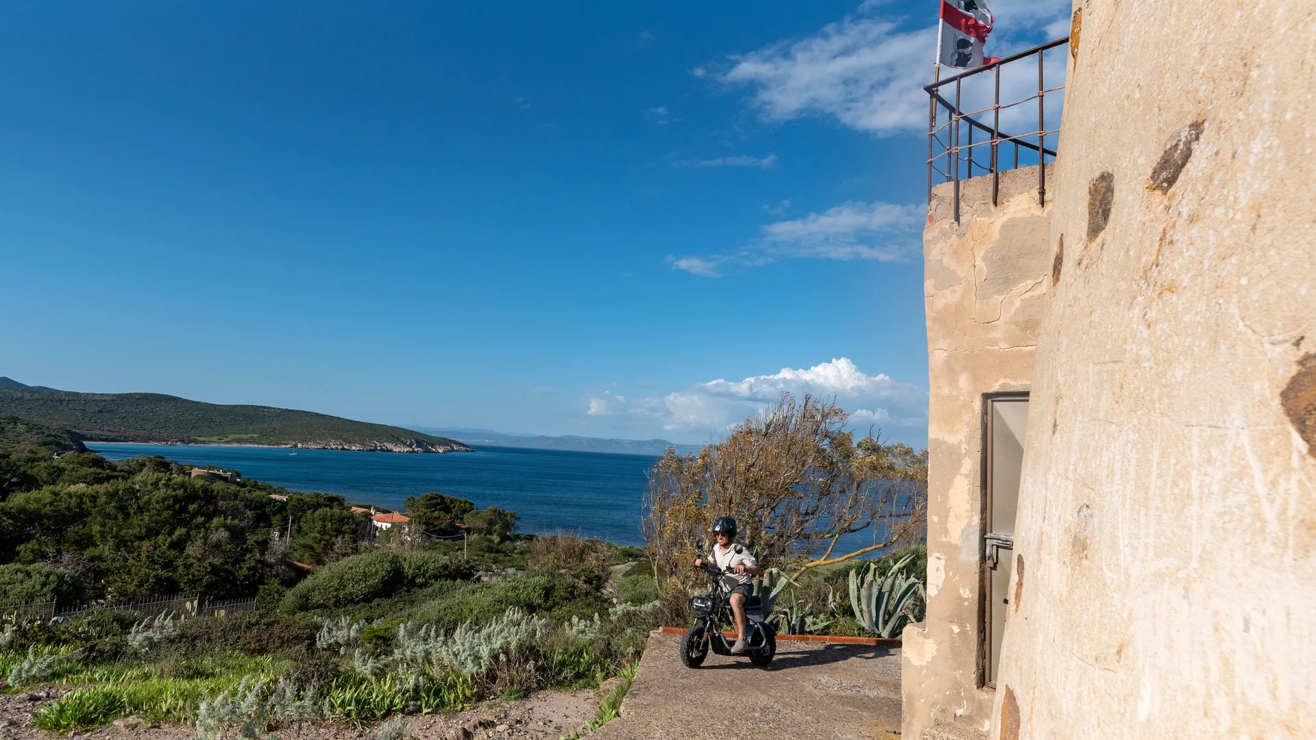 Person mit Helm auf E-Roller neben Turm mit Flagge und Meerblick