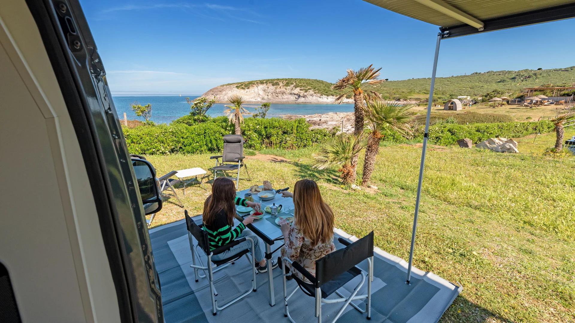Zwei Mädchen essen draußen mit Blick auf Meer und grüne Küste.
