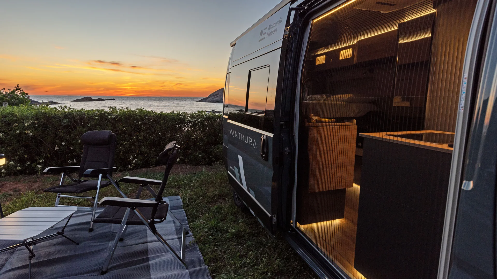 Wohnmobil mit offenen Vorhängen und Stühlen, Sonnenuntergang am Meer im Hintergrund
