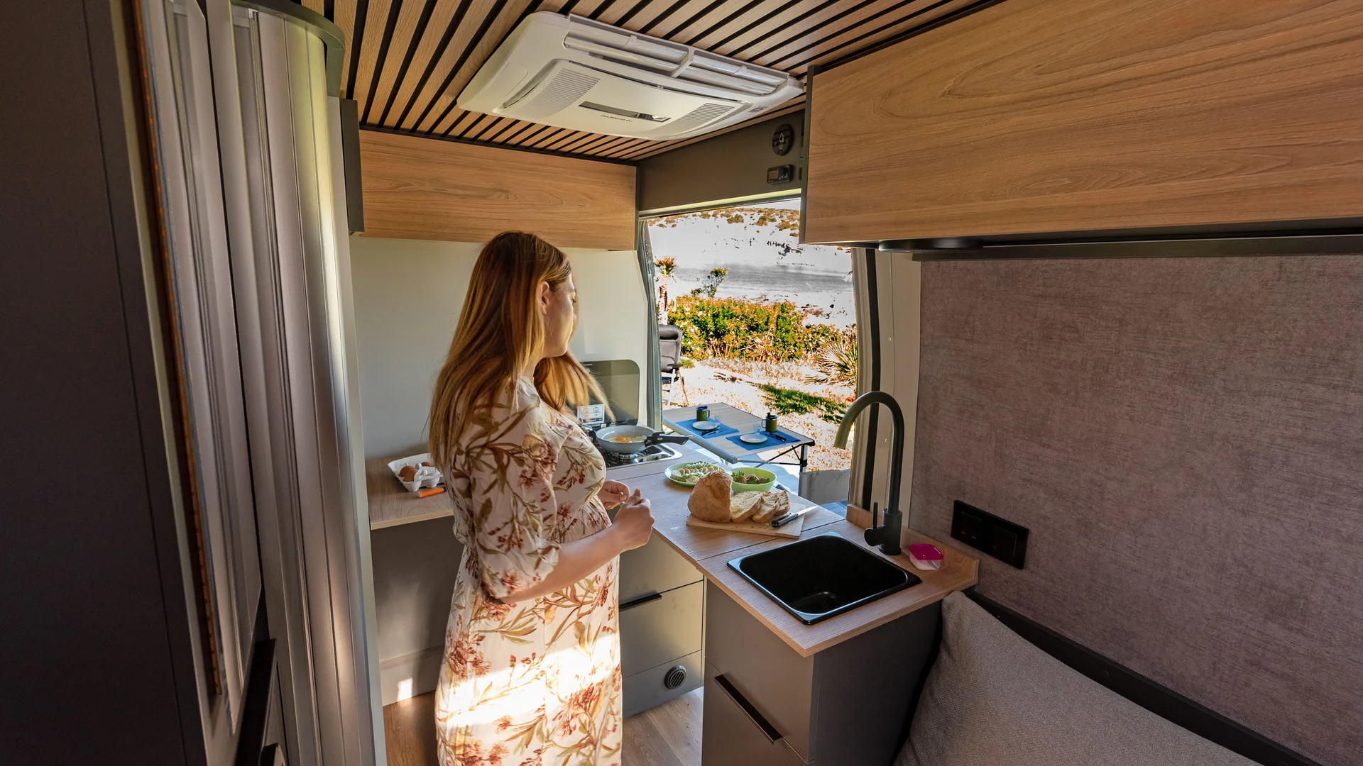 Frau kocht in kleinem Wohnmobil mit Holzmöbeln und Außenausblick
