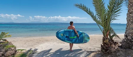 Mann mit blauem Surfbrett am Strand vor dem Meer mit Palmen