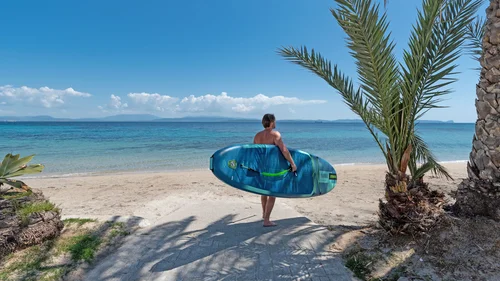 Mann mit blauem Surfbrett am Strand vor dem Meer mit Palmen