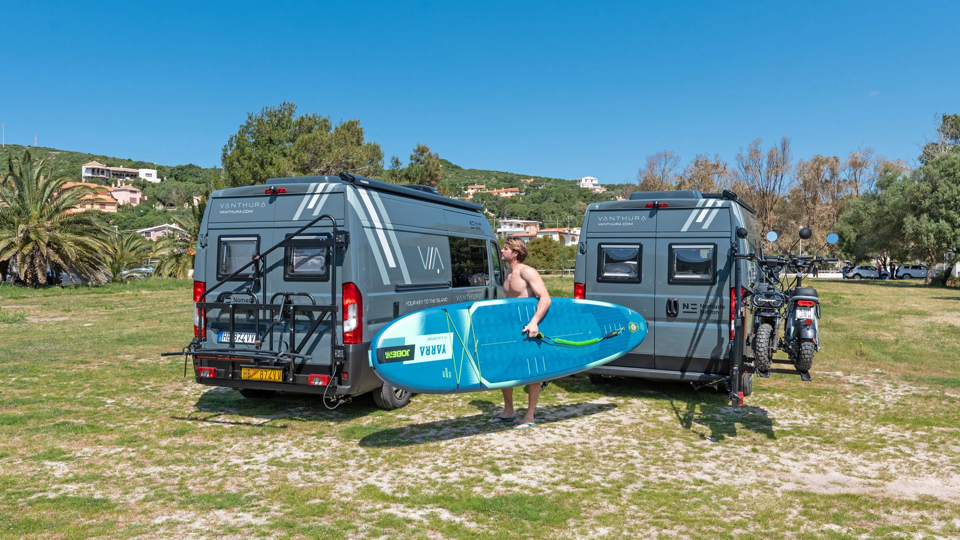 Zwei graue Wohnmobile mit Mann, der blaues Surfbrett bei Sonne trägt